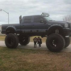 two girls standing in front of a monster truck on the side of a road with it's huge tires