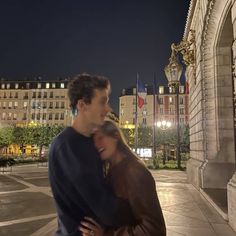 a man and woman standing in front of a building at night with the lights on