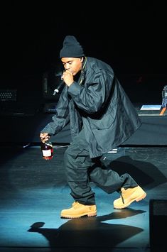 a man in black jacket and yellow boots on stage holding a bottle with both hands