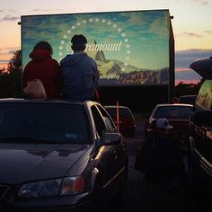 two people sitting on the back of a car in front of a movie screen at dusk