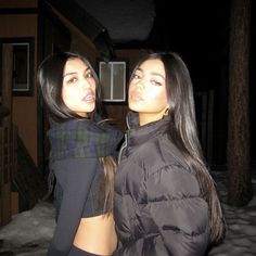 two young women standing next to each other in the snow at night, one is wearing a black jacket