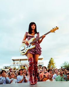 a woman in red boots holding a white guitar and standing on stage with people behind her