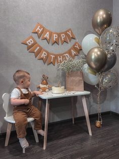 a little boy sitting at a table with a birthday cake and balloons in the background