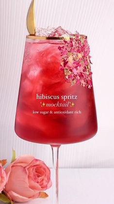 a red cocktail with pink flowers and gold garnish on the rim next to a white wall