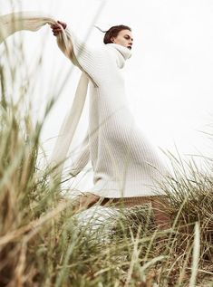 a woman standing in tall grass wearing a white sweater and scarf with her arms outstretched