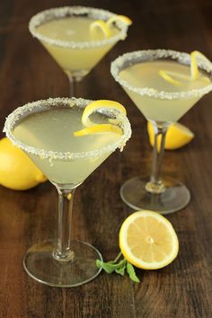 three martinis with lemon garnish sit on a wooden table next to sliced lemons