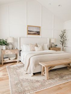 a bedroom with white walls and wood flooring has a large bed, two nightstands, and a bench in front of the bed