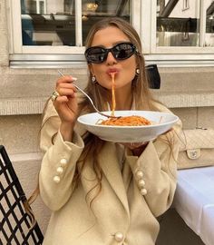 a woman in sunglasses is eating spaghetti from a white bowl with a fork and spoon