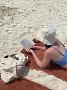 a woman reading a book while laying on a towel in the sand at the beach