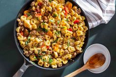 a skillet filled with pasta and sausage on top of a table next to a wooden spoon