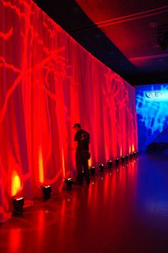 two people standing in front of a red curtain with blue lights on the wall behind them