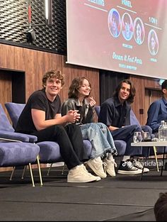 four people sitting on blue chairs in front of a projector screen at an event