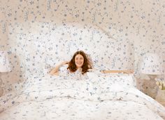 a woman laying on top of a white bed in a room with blue floral wallpaper