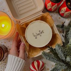 a person holding a container with the word winter written on it next to christmas decorations and candles