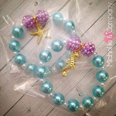 two little mermaid bracelets with blue pearls and gold charms on them, sitting in a clear plastic bag