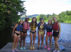 four girls in bathing suits standing on a dock by the water with life vests