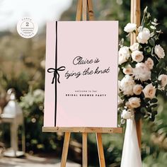 an easel with a pink sign and white flowers on it, next to a floral arrangement