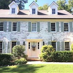 a large white brick house with shutters on the front and side windows, surrounded by greenery