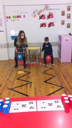 two children playing with toys on the floor in a school room, one is sitting at a table and the other is standing