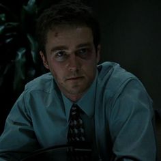 a man wearing a shirt and tie sitting at a table in the dark with his hands on his hips