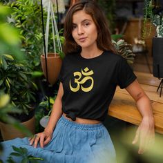 a woman sitting on a bench wearing a black shirt with gold omen symbol in front of potted plants