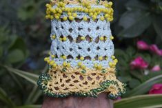 a hand holding a crocheted object in front of flowers