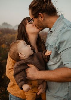 a man and woman kissing their baby boy in the middle of an outdoor photo shoot