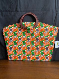 Vibrantly Colored Ankara Handbag has oval shaped wooden handles with a snap closure. Handmade 100% cotton Multicolor Top Handle Bag With Rolled Handles, Cotton Tote Bag With Detachable Handle, Orange Cotton Shopping Bag, Cotton Top Handle Bag With Leather Handles, Rectangular Cotton Beach Bag With Leather Handles, Top Handle Cotton Bag With Leather Handles, Multicolor Bag With Round Handle For Daily Use, Multicolor Bags With Round Handle For Daily Use, Multicolor Tote Bag With Bamboo Handle