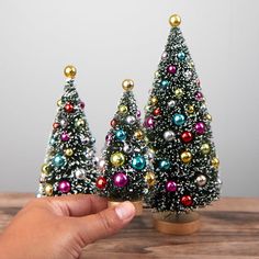 a hand holding a tiny christmas tree in front of two small trees on a wooden table