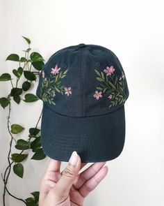 a person holding up a baseball cap with embroidered flowers on the front and side, in front of a plant