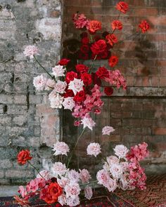 red and pink flowers are in a vase on a rug next to a brick wall