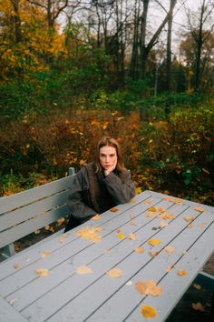 a woman sitting at a wooden table with leaves on the ground in front of her