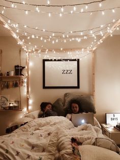 two people are laying in bed with lights strung above them and one person is using a laptop