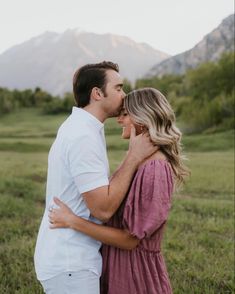 a man and woman standing in the grass with mountains in the background