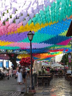 many colorful umbrellas hanging from the ceiling in an outdoor market area with people walking under them