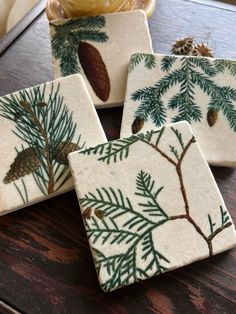 four ceramic coasters with pine cones and evergreen needles on them, sitting on a table