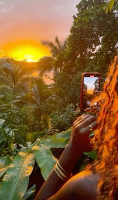 a woman taking a photo with her cell phone in front of the sun setting over some trees