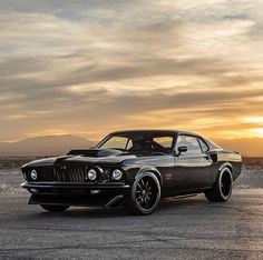 a black muscle car parked in an empty lot with the sun setting behind it and clouds