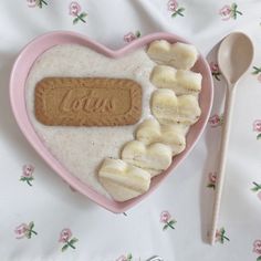 a pink heart shaped bowl filled with oatmeal and banana slices next to a spoon