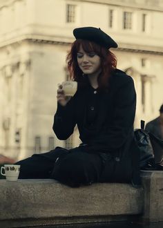 a woman with red hair sitting on a ledge drinking from a cup