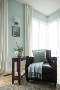 a living room with a chair, table and two large windows in it's corner