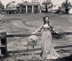 an old photo of a woman leaning on a fence in front of a large house