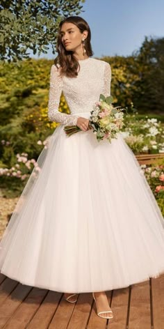 a woman in a white wedding dress standing on a wooden deck with flowers and greenery