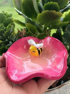 a hand holding a pink flower with a yellow bee on it's head in front of a potted plant