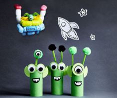 three green toys with eyes and mouths are lined up in front of a space rocket