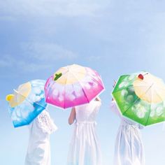 three women in white dresses are holding umbrellas on the beach and looking at the sky