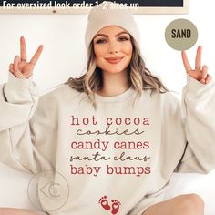 a woman sitting on the floor with her hands in the air and wearing a sweatshirt that says hot cocoa cookies, candy canes, santa claus baby bumps