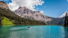 two people in a small boat on a lake surrounded by mountains and pine trees under a blue sky with white clouds