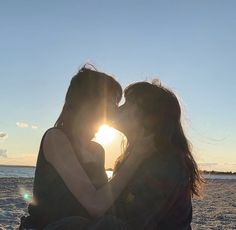 two women sitting on the beach kissing each other with the sun setting in the background