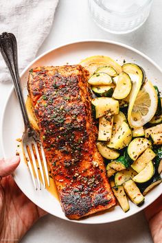 someone is holding a plate with salmon, zucchini and squash on it next to a fork